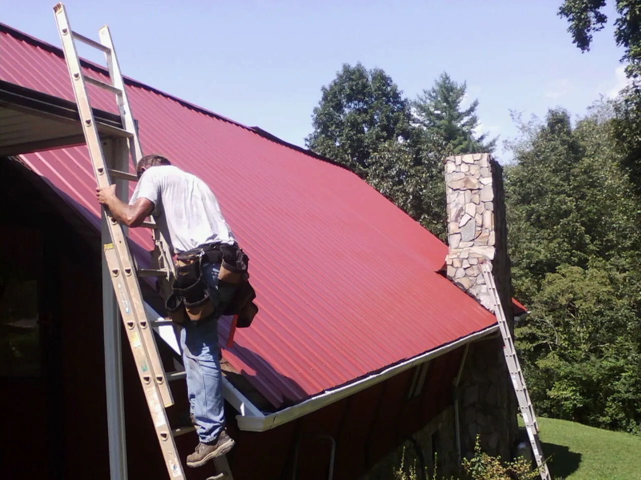 Roofer on a ladder beside a red metal roof and stone chimney — Soffit & Fascia Repair by Roofing Brothers in Livonia